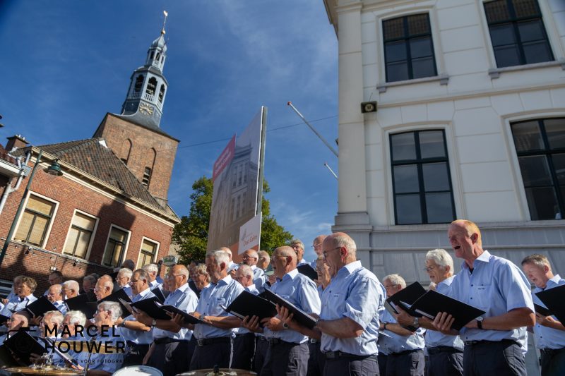 Koorleden van Mannenzangvereniging Inter Nos zingen op de Markt in Groenlo tijdens een zomerconcert, met de toren van de Oude Calixtuskerk op de achtergrond.