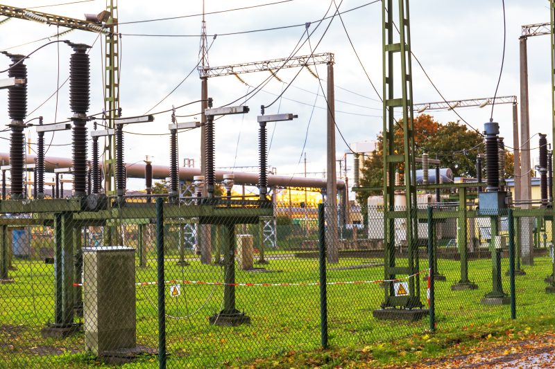An electrical substation with various power equipment, including transformers and wires, surrounded by a fence and green grass.